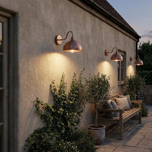Three copper-toned outdoor wall sconces mounted on a stucco house wall, casting warm light onto a stone patio beside a wooden bench and potted plants at dusk.