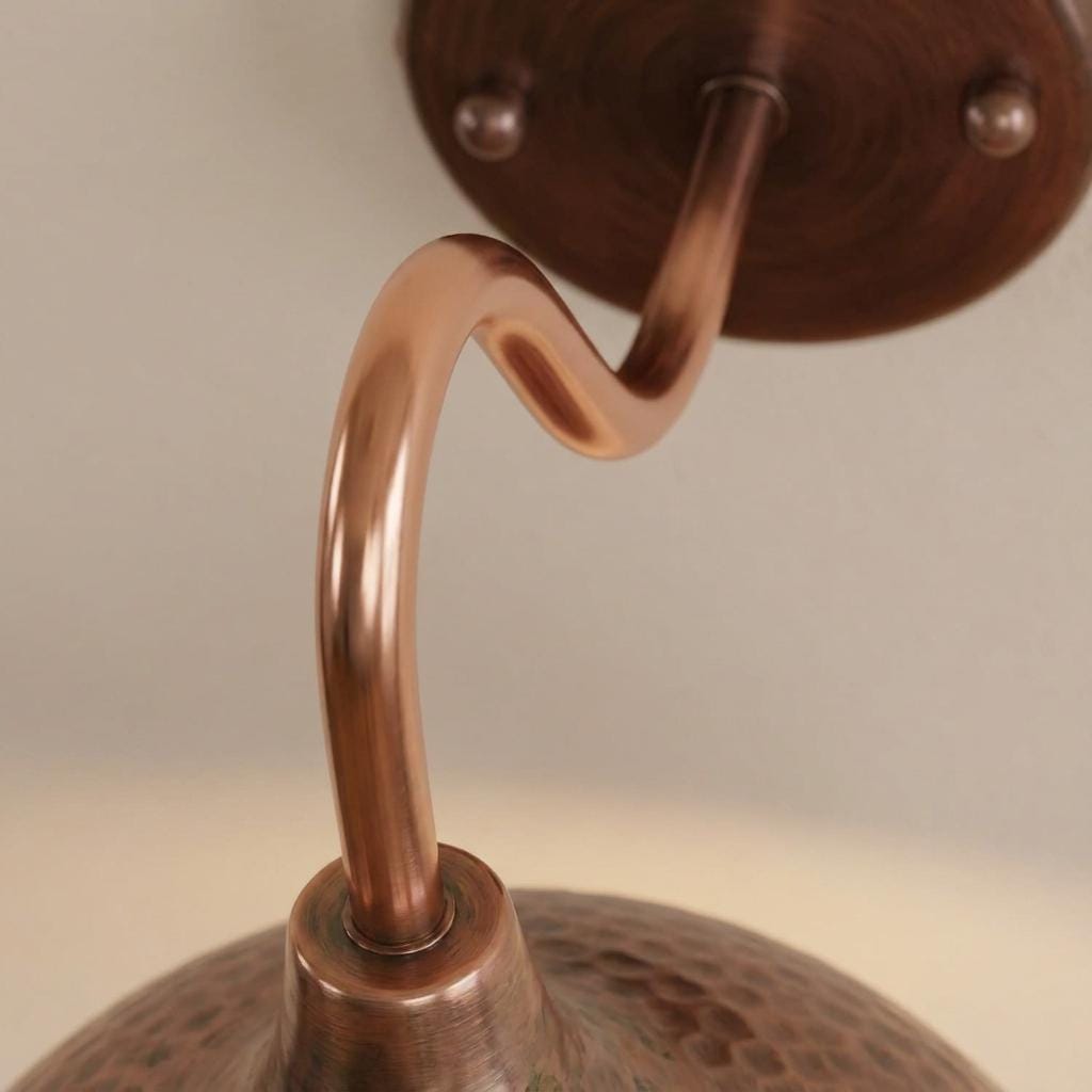 Close-up of a copper-toned metal wall fixture with a curved hook-shaped stem, textured lower section, and round mounting plate against a plain light background.