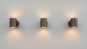 Three matching cylindrical wall sconces with a distressed bronze and turquoise patina finish, mounted on a plain light gray wall and casting warm light downward.