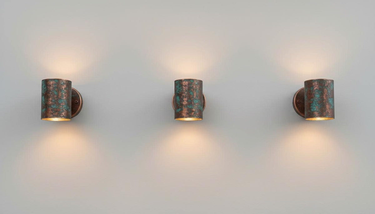 Three matching cylindrical wall sconces with a distressed bronze and turquoise patina finish, mounted on a plain light gray wall and casting warm light downward.