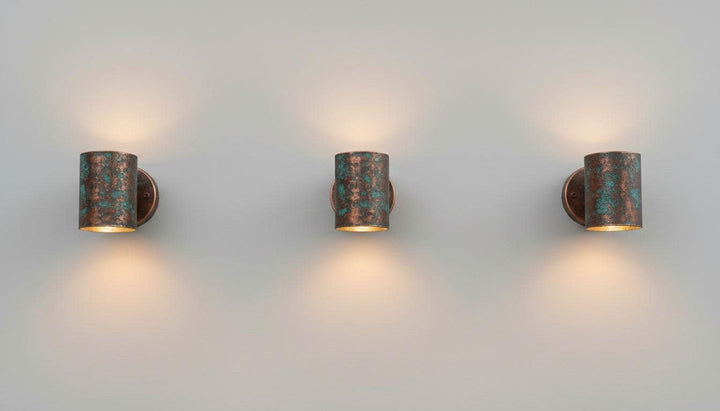 Three matching cylindrical wall sconces with a distressed bronze and turquoise patina finish, mounted on a plain light gray wall and casting warm light downward.