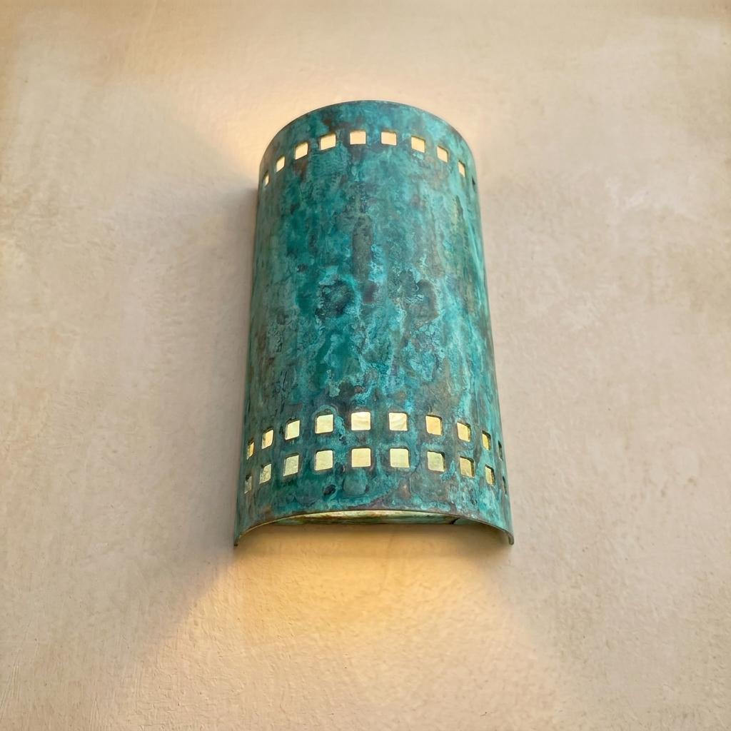 A cylindrical wall sconce with a distressed turquoise and green patina finish, featuring square cutout details on the sides that cast warm light onto the beige wall behind it.