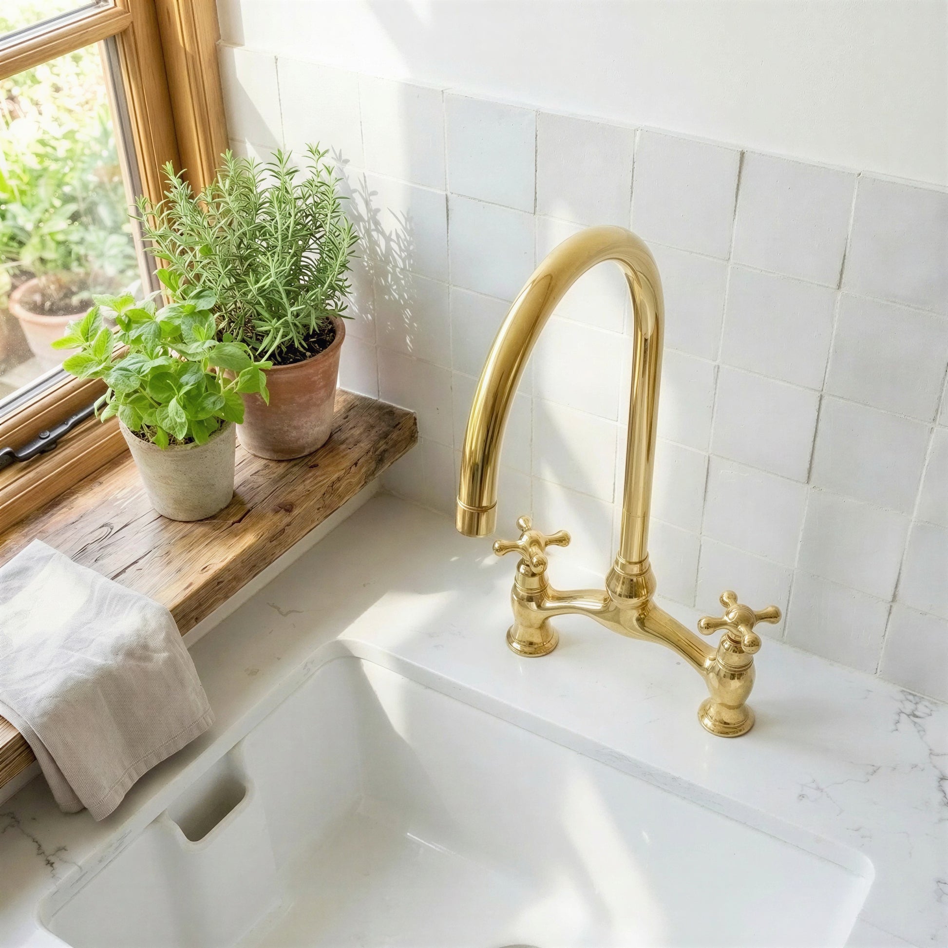 A polished brass bridge faucet with two cross handles installed in a white farmhouse sink, set against a white subway tile backsplash with a wooden window sill holding potted green plants.