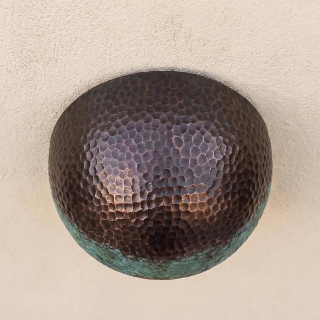 A round hammered metal wall sconce with a dark bronze finish and a greenish patina along the lower edge, mounted on a textured beige wall.