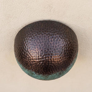 A round hammered metal wall sconce with a dark bronze finish and a greenish patina along the lower edge, mounted on a textured beige wall.