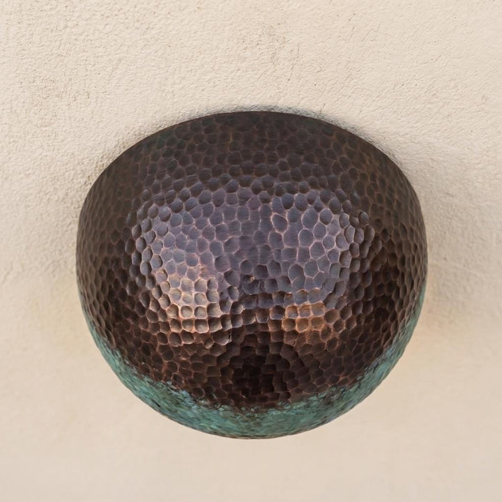 A round hammered metal wall sconce with a dark bronze finish and a greenish patina along the lower edge, mounted on a textured beige wall.