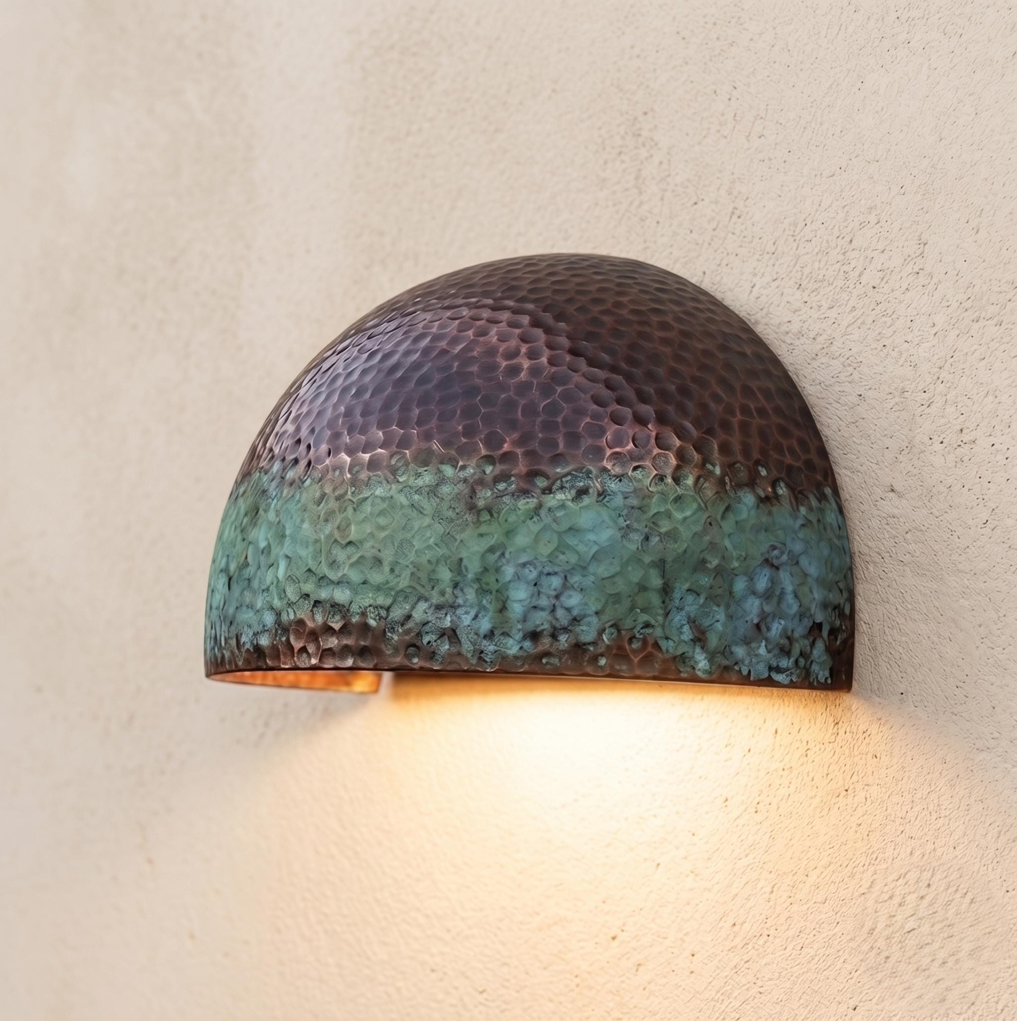 A hammered copper dome wall sconce with a dark bronze top half and a green-blue patina lower half, mounted on a textured beige wall and casting warm light downward.