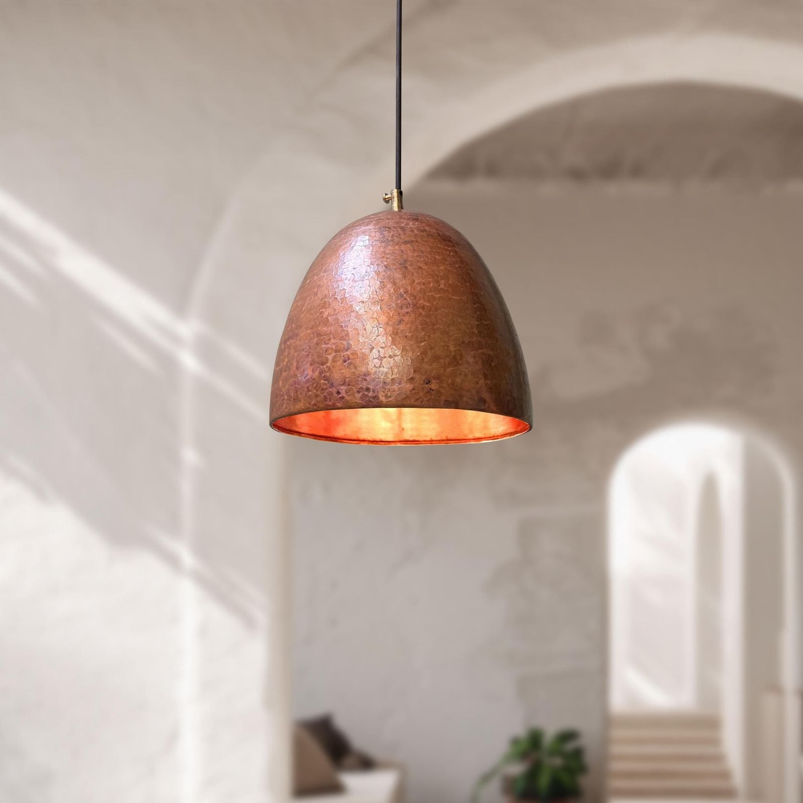A dome-shaped pendant light with a textured copper-colored metal shade and a warm orange interior, hanging in a softly blurred architectural interior with arches and sunlight.